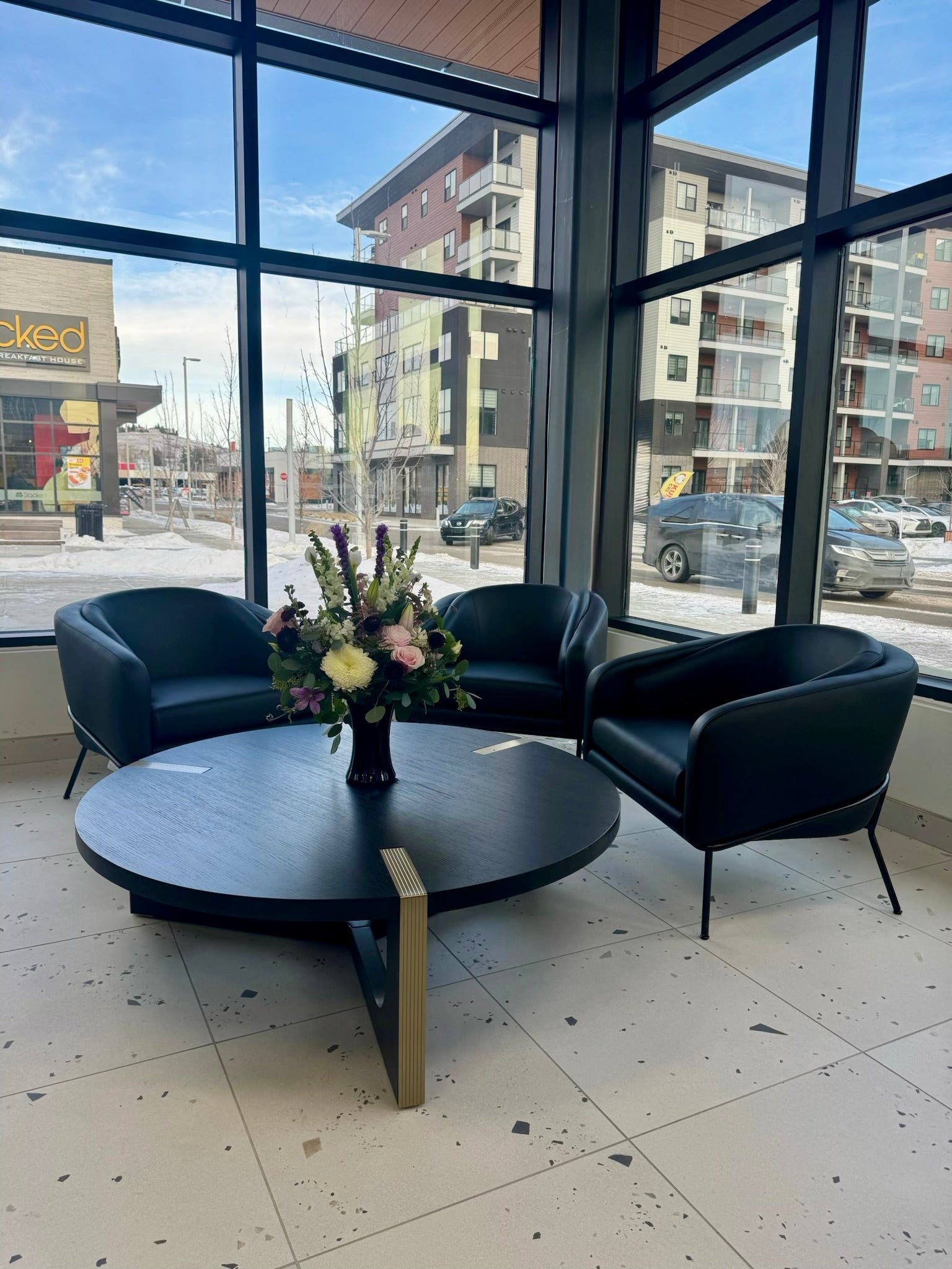 Modern interior with black chairs and a round table in front of large windows showing an urban street view. Curious Salons Northland Village Shops
