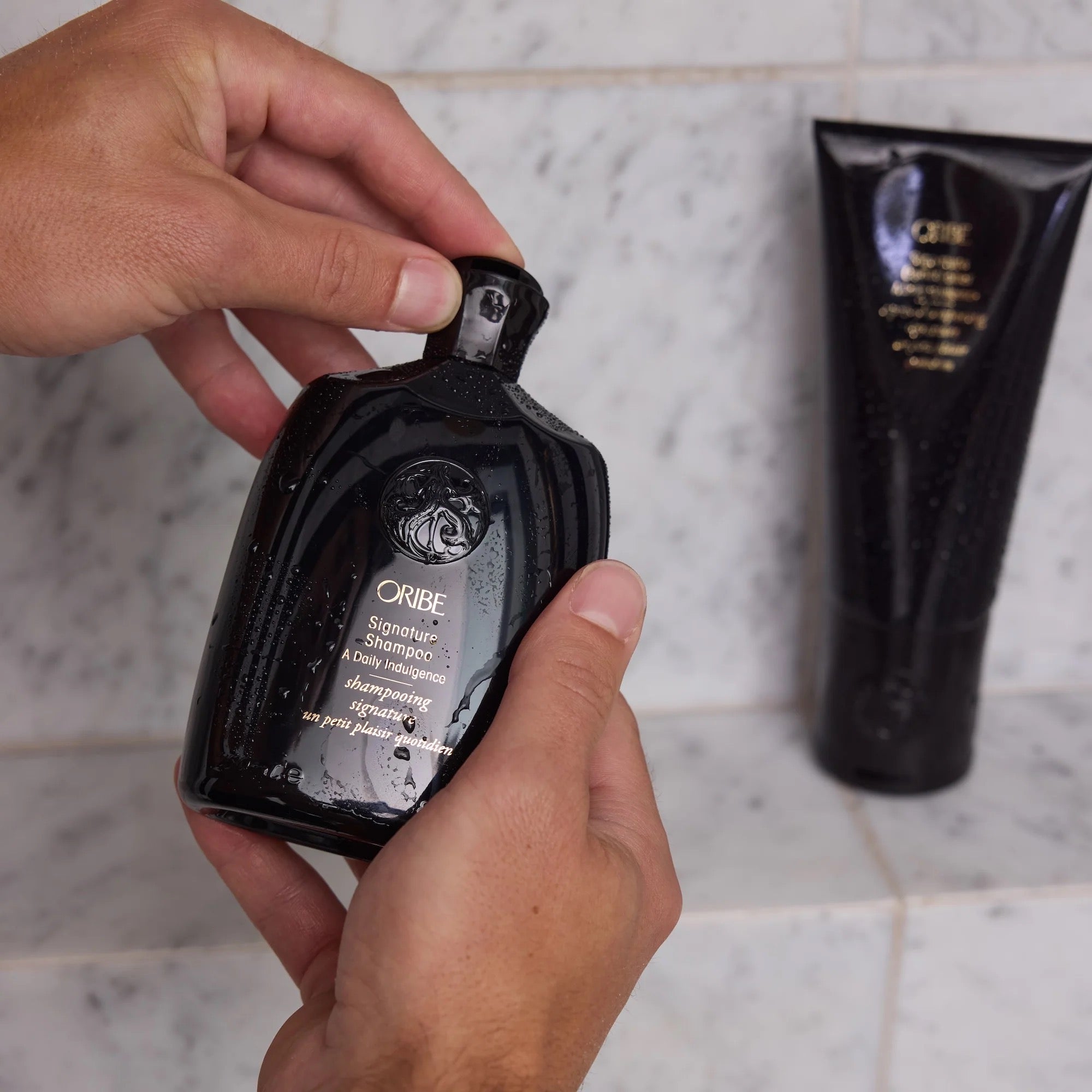 Person holding Oribe shampoo bottle with a tube of product in the background Oribe Signature Shampoo luxury daily shampoo for all hair types in Canada