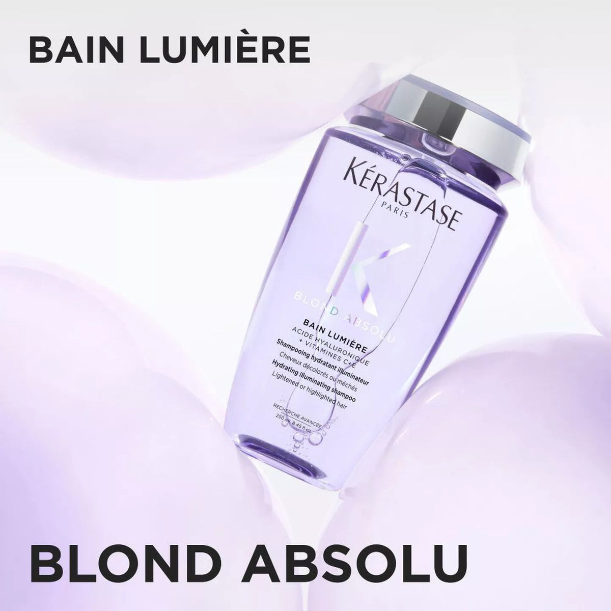 Kérastase Bain Lumière Blond Absolu hair developer bottle in a clear rectangular container with a silver cap, set against a soft white and pale lavender background with decorative shapes. Large text reads 'BAIN LUMIÈRE' at the top and 'BLOND ABSOLU' at the bottom.