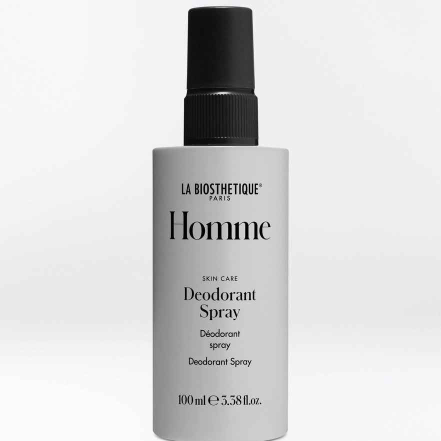 A 100ml bottle of La Biothetique Homme Deodorant Spray on a light gray background. The bottle is a light gray color with a black spray nozzle and black text on the label.