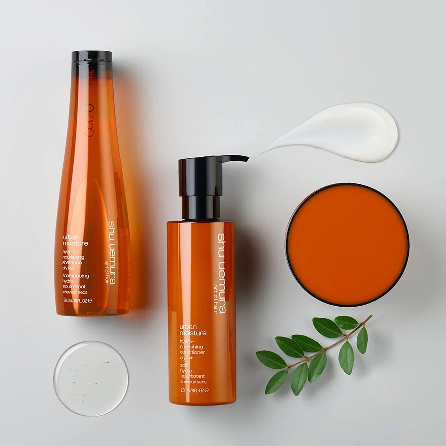 Set of orange skincare Shu Uemura Urban MoistureHydrate Dry Hair Canada
products with pump bottle, jar, and leaves on a light background