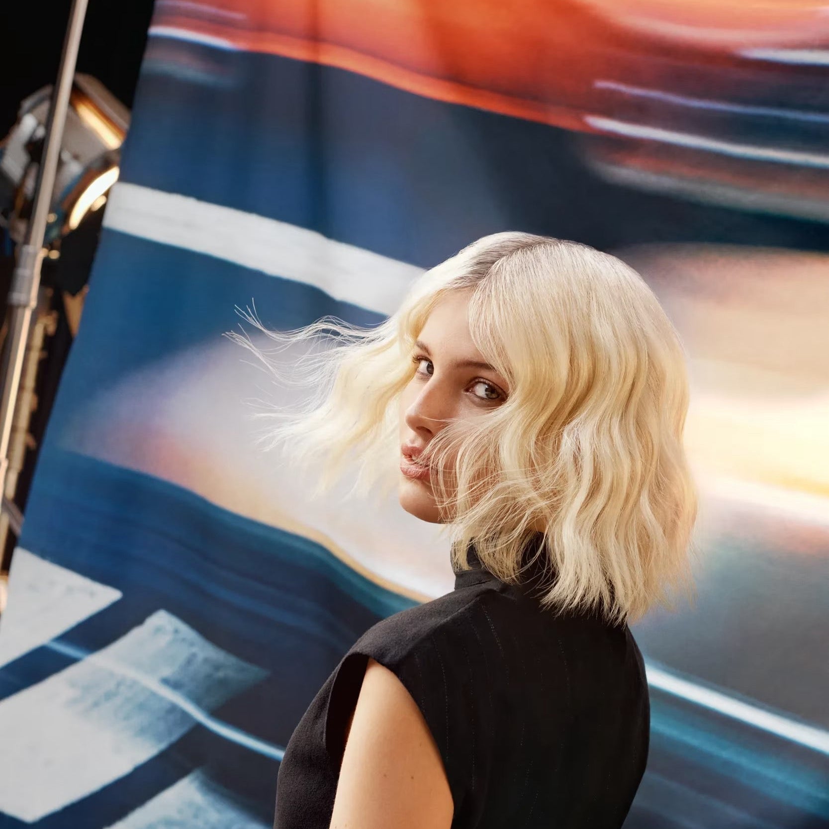 Woman with blonde hair and a black dress standing in front of a colorful abstract painting.