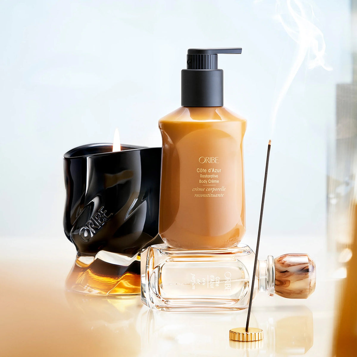 Oribe Cote d'Azur body care set featuring a brown bottle with a black pump, a black scented candle, and a glass incense holder with a smoking incense stick, arranged on a reflective surface.