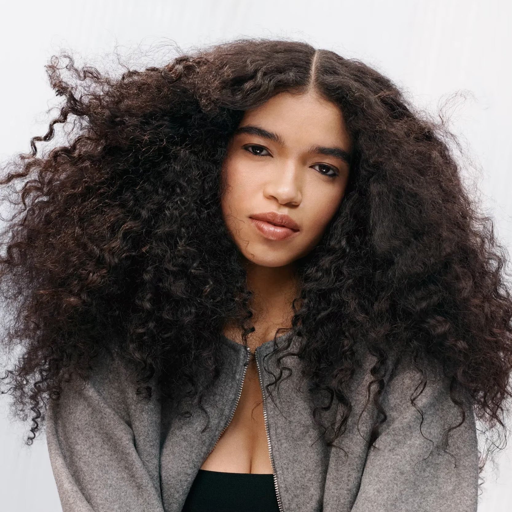 Woman with voluminous curly hair wearing a gray cardigan over a black top against a white background