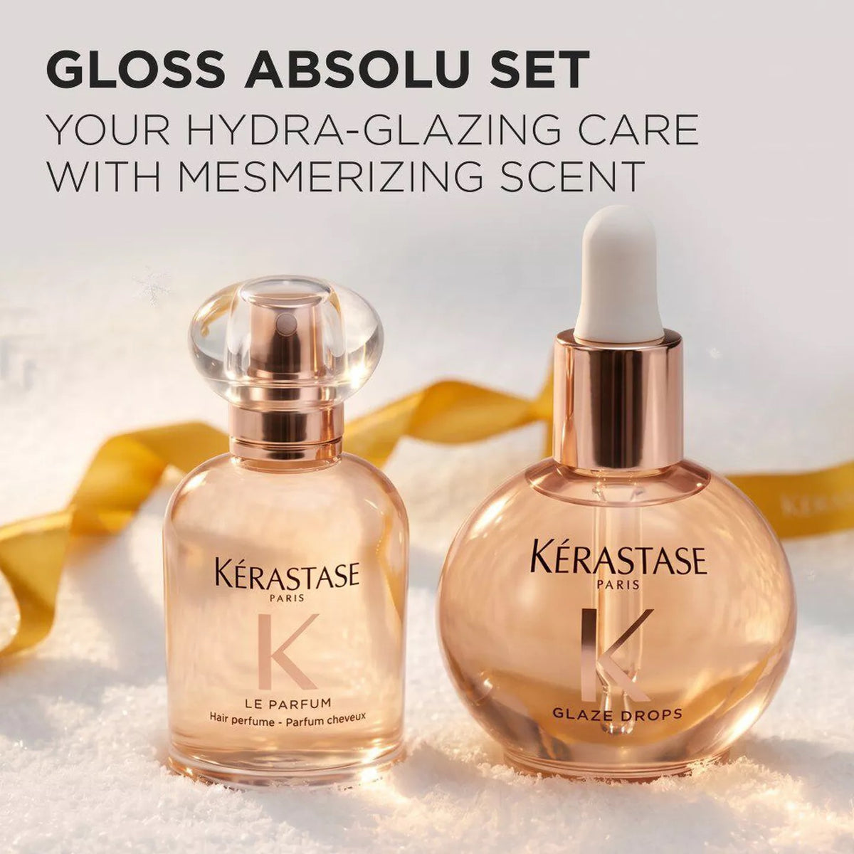 Two Kérastase Paris hair care bottles from the Gloss Absolu Set, featuring Le Parfum and Glaze Drops, styled on a snowy surface with a gold ribbon and a soft light background.