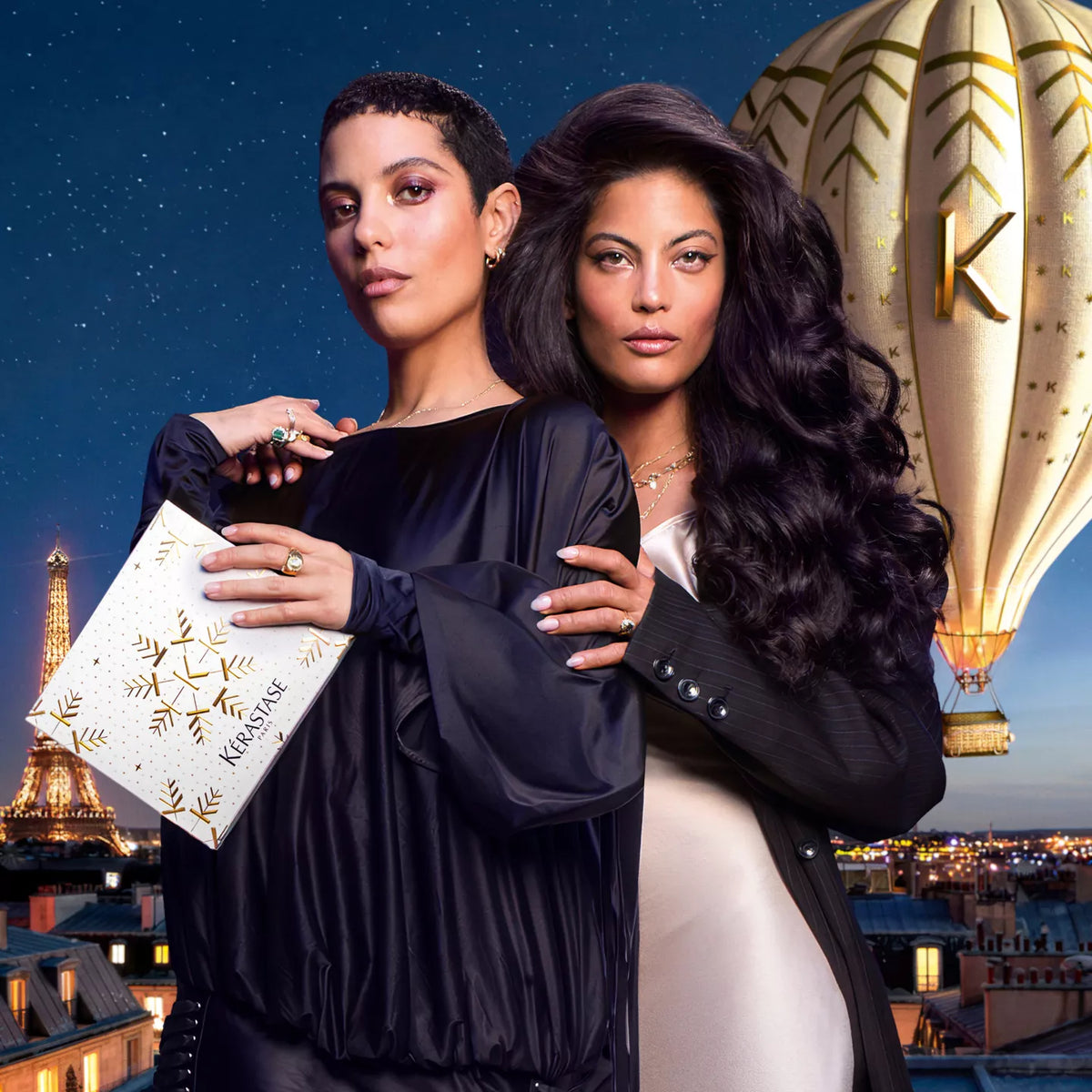 Two women posing together in a Parisian-themed setting at dusk, with the Eiffel Tower and a hot air balloon in the background. Kerastase Holiday Packs 2026 Curious Salons