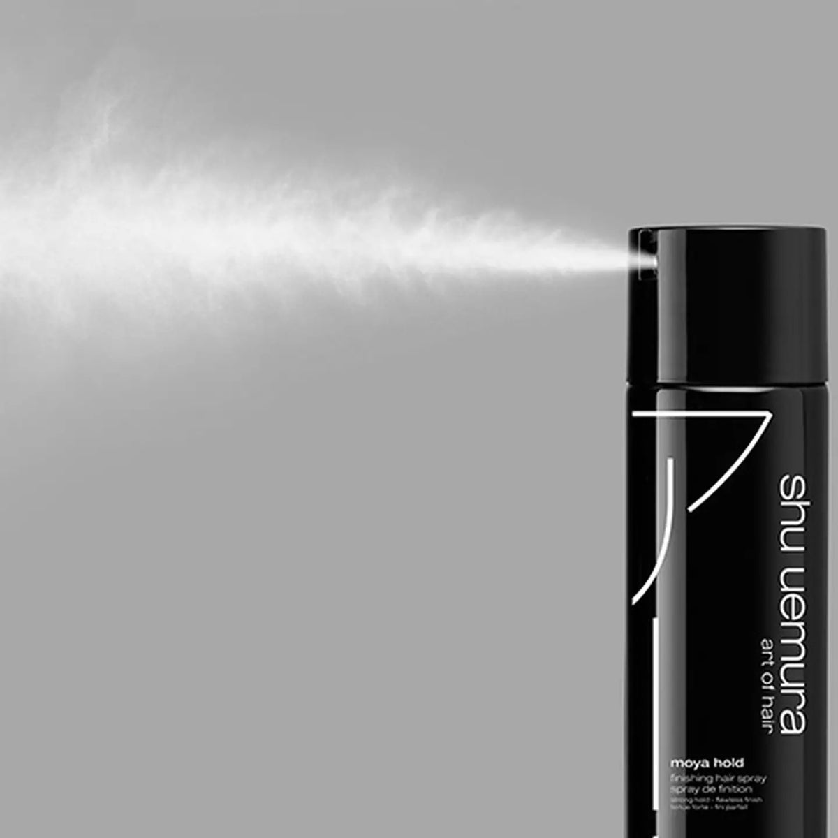 Shu Uemura Moya Hold Hair Sprinkle Fixing Spray bottle on the right side, with a fine mist of white spray extending to the left against a solid gray background.