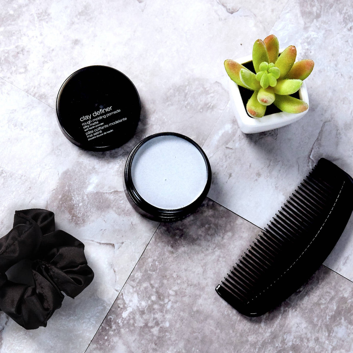 Hair care products including a jar, comb, and scrunchie on a textured surface with a small plant. Curious Salons
