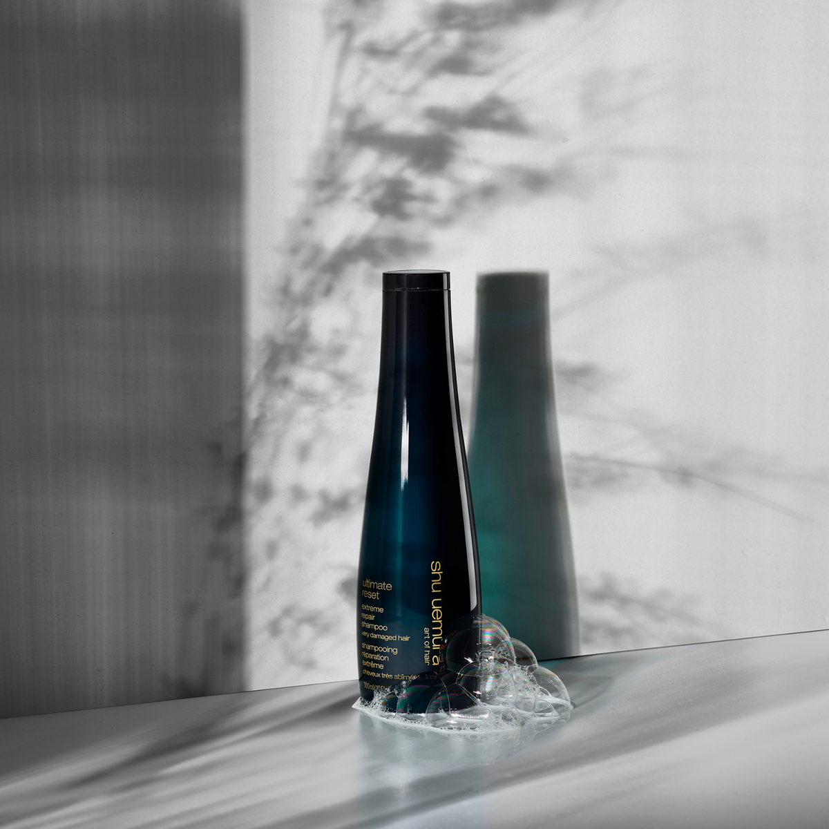 Shu Uemura Ultimate Reset bottle on a reflective surface with a blurred background and  drop of shampoo
