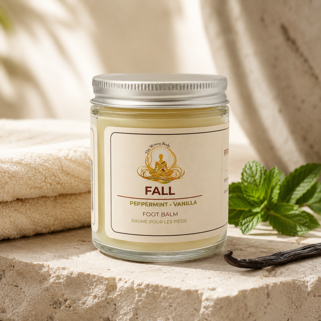 A glass jar of fall foot balm with a silver lid and cream label, styled on a stone surface with a folded towel, mint leaves, and a vanilla bean. The Woven Body Peppermint Vanilla Foot Balm  Clean Product